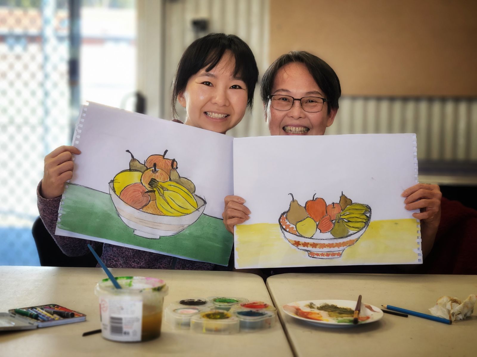 Two women holding up their paintings of fruit and smiling.