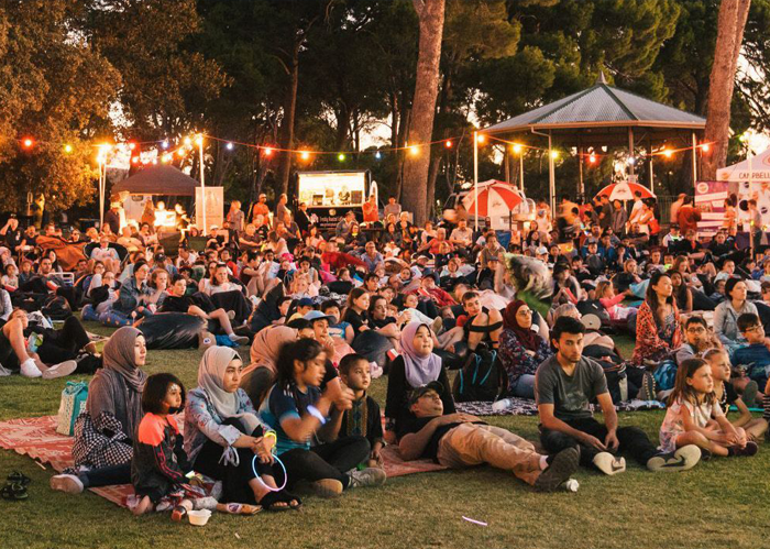 A large group of people sitting in a park at dusk watching an outdoor movie.
